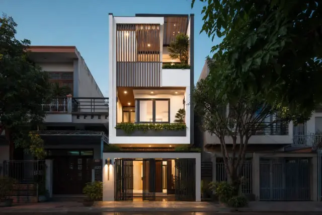 Mặt tiền nhà ống 3 tầng hiện đại với tường trắng, lam gỗ dọc và ban công cây xanh lúc chiều tối