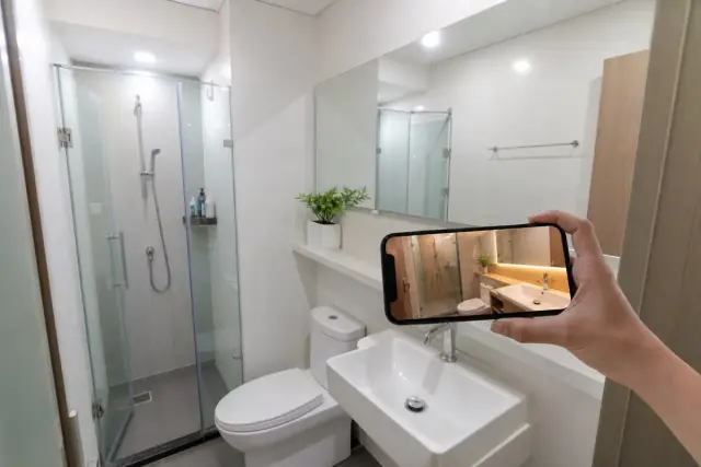 Phòng tắm chung cư nhỏ với gạch trắng, kính cường lực vòi sen và người xem phối cảnh WC do AI thiết kế trên điện thoại