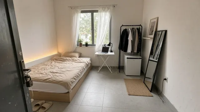Phòng trọ 20m2 tường trắng, sàn gạch sáng với giường thấp, bàn gấp cạnh cửa sổ, kệ treo quần áo đứng, gương đứng và đèn LED ấm sau đầu giường