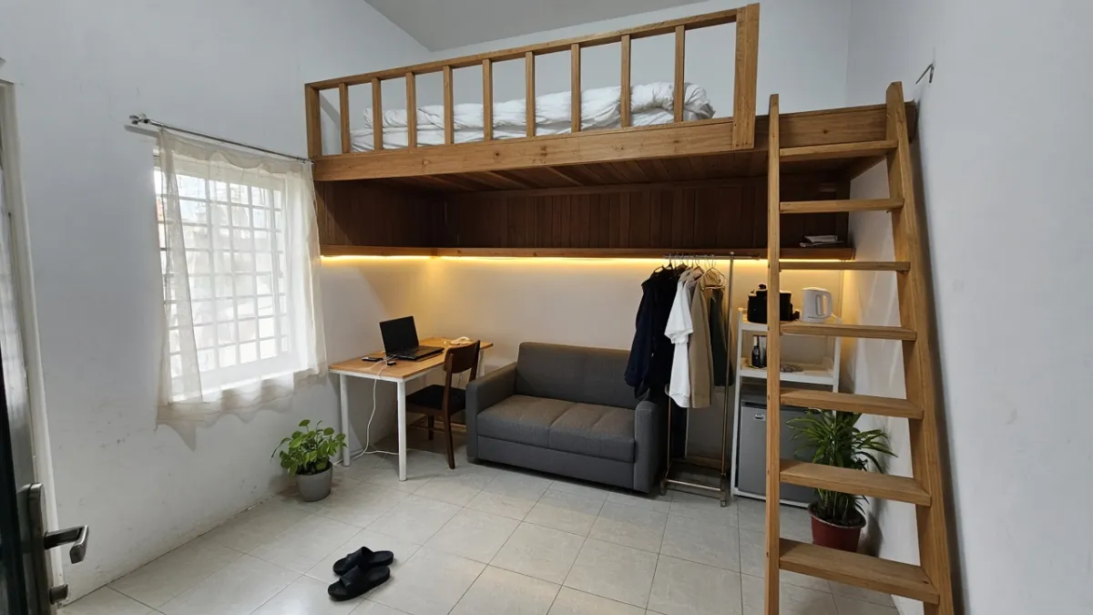 Phòng trọ có gác lửng gỗ, phía dưới gác đặt bàn làm việc, sofa xám nhỏ, kệ treo quần áo và tủ lạnh mini, đèn LED ấm gắn dưới mép gác