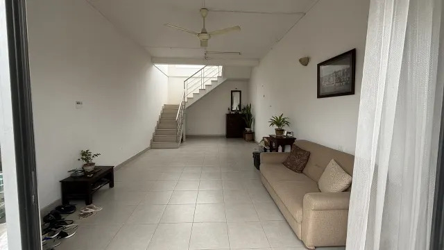 Nhà ống nhìn từ cửa trước, sofa dọc tường phải, cầu thang bê tông sát tường trái và ô sáng phía trên thang