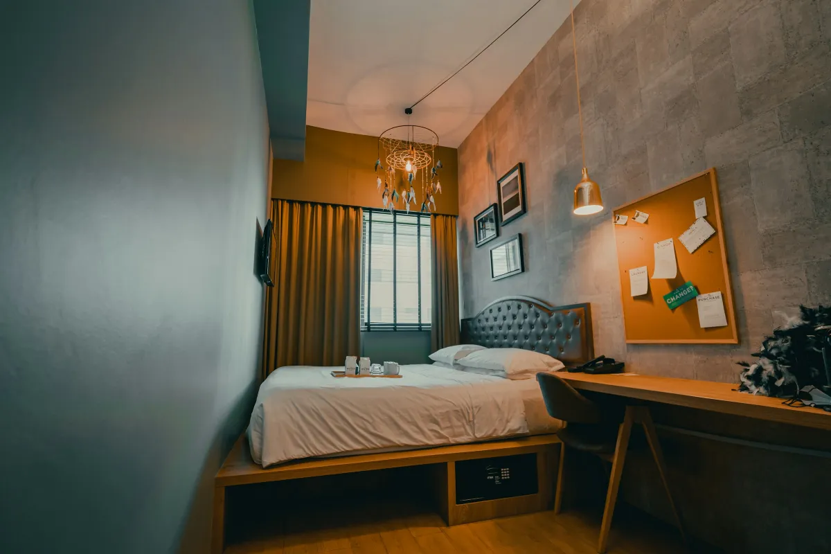 Phòng ngủ hẹp dài với giường đặt trên bục gỗ có hộc chứa đồ, bàn làm việc gỗ chạy dọc tường bên phải và cửa sổ cuối phòng